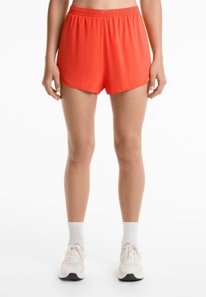 Pantaloni sportivi rossi con cintura elastica, realizzati in un tessuto leggero e liscio, dal taglio morbido e con orli arrotondati. Abbinati a scarpe da ginnastica leggere e calzini.