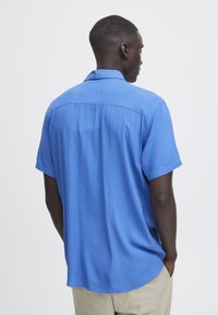 Camisa de manga corta en azul brillante, confeccionada en material ligero. Presenta un corte relajado con cuello estándar y detalle de pliegues en la parte posterior.