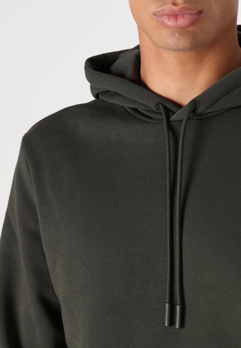 Haut à capuche vert foncé avec un cordon de serrage, présentant une texture côtelée et une coupe décontractée. Tissu lisse et design minimaliste.