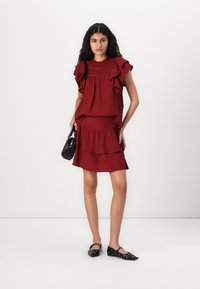 Red ruffled dress with smocked bodice, layered skirt, and a black handbag. Model wears black flats with circular accents. Simple backdrop.