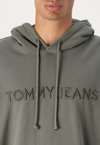 Felpa con cappuccio grigia con la scritta 'TOMMY JEANS' ricamata in grigio scuro. Ha una texture morbida e lacci regolabili sul cappuccio.