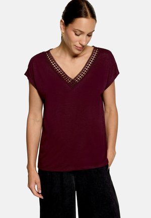 Femme portant un haut violet foncé à manches courtes avec un détail en dentelle en forme de V et un pantalon noir, debout avec une main dans la poche.