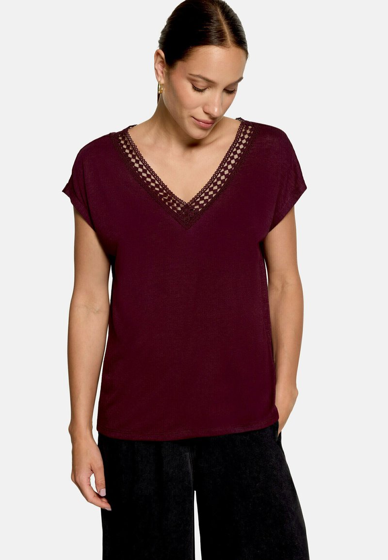 Femme portant un haut violet foncé à manches courtes avec un détail en dentelle en forme de V et un pantalon noir, debout avec une main dans la poche.