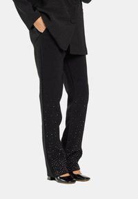 Pantalones negros de corte recto, decorados con pequeños adornos brillantes cerca del dobladillo, combinados con zapatos negros de punta.