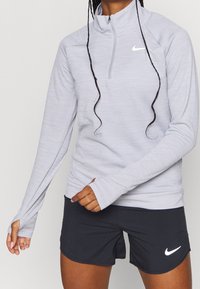 Maglione grigio chiaro a maniche lunghe con zip a un quarto e aperture per il pollice, realizzato in tessuto testurizzato, con il logo Nike bianco sul petto, abbinato a pantaloni corti neri.