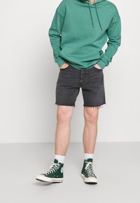 Henkilö, joka käyttää vihreää hupparia ja mustia farkkushortseja, joissa on harventuneet helmat, seisoo kädet taskuissa, vihreät korkeavartiset sneakersit ja valkoiset sukat.