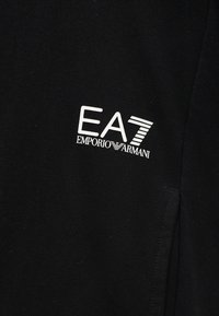 Μαύρο ύφασμα με λευκό λογότυπο "EA7 Emporio Armani" τυπωμένο στην αριστερή πλευρά του στήθους ενός ενδύματος.