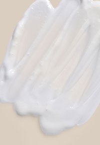 Vit krämig substans med en slät, glansig textur, applicerad i ett vågigt mönster på en ljus beige bakgrund.
