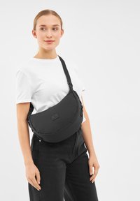 Borsa a spalla in tessuto nero con forma curva, tracolla regolabile e parte anteriore piatta con un piccolo logo rettangolare.