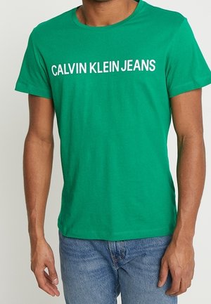 Zielona bawełniana koszulka z białym nadrukiem "CALVIN KLEIN JEANS" na wysokości klatki piersiowej, z okrągłym dekoltem i krótkimi rękawami. Nosi się ją z niebieskimi dżinsami.