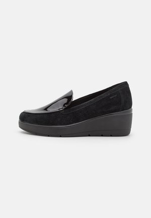 Zapatos negros de estilo slip-on con suela de cuña, que presentan una parte delantera de charol y un cuerpo de gamuza suave, con textura lisa y forma redondeada.