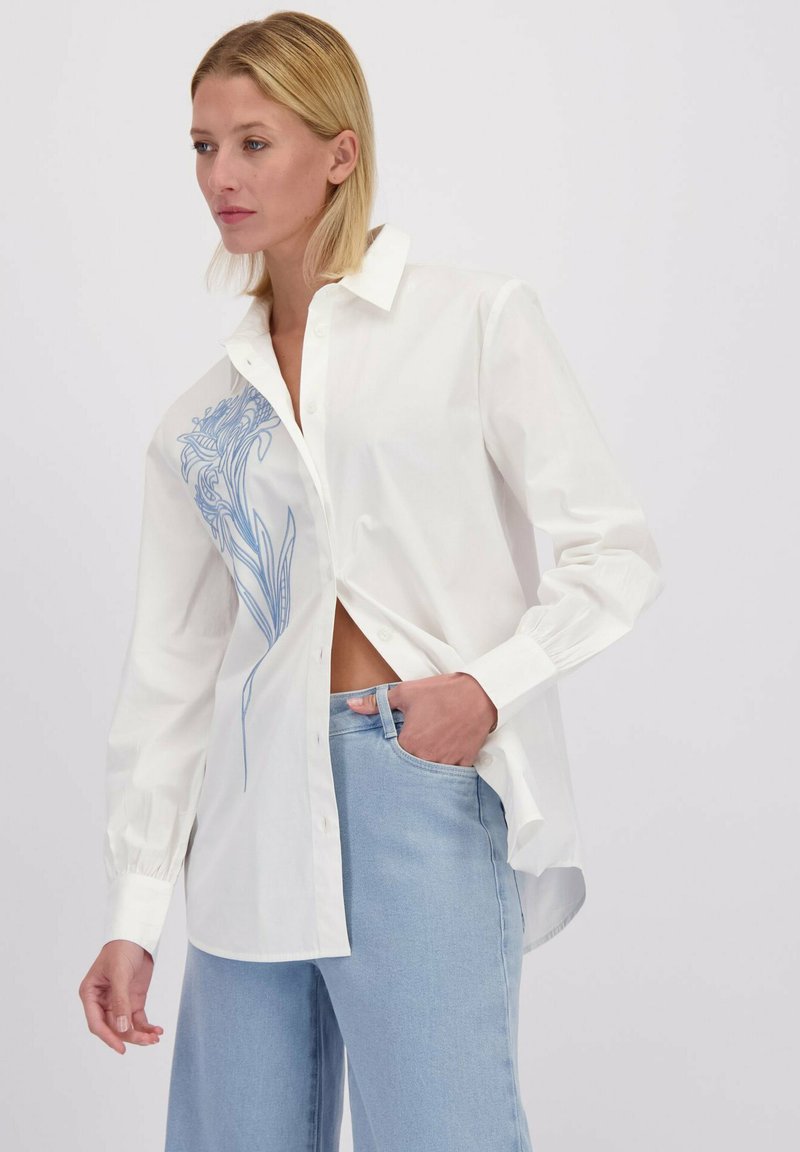 Vrouw in een witte button-up shirt met blauwe bloemenborduurwerk aan de linkerkant en lichtblauwe spijkerbroek, staand tegen een effen achtergrond.