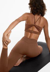 Soutien-gorge de sport marron avec un design à dos torsadé, associé à un legging taille haute assorti, le tout en matériau lisse et extensible. Tapis de yoga noir en dessous.