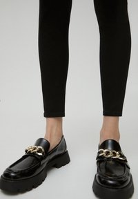 Mocasines de charol negro con suela gruesa, que presentan un acento de cadena dorada en el empeine y una textura suave y brillante.
