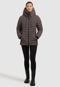 Donna che indossa una giacca a vento marrone scuro con colletto alto, pantaloni aderenti neri e stivaletti neri, in piedi su uno sfondo grigio uniforme.