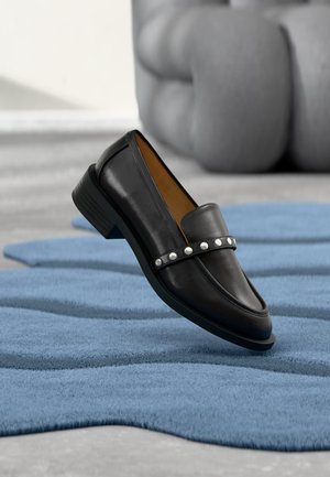Mocassin en cuir noir avec des clous argentés sur la bride flottant au-dessus d'un tapis bleu texturé et d'un mobilier gris rembourré en arrière-plan.