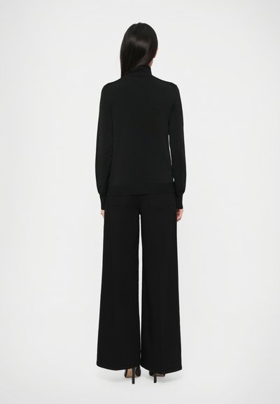 Kvinde står med ryggen til iført en sort langærmet turtleneck og brede sorte bukser med højhælede sko på en hvid baggrund.