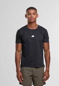 Svalnatý muž stojící čelem dopředu, oblečený v černém tričku s krátkým rukávem a bílým logem a olivově zelených šortkách na jednobarevném pozadí.