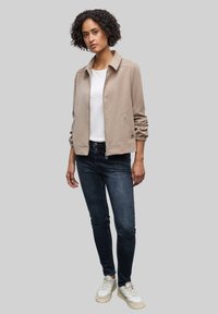 Beżowa kurtka bomberka z kołnierzykiem, zamkiem z przodu, dwoma bocznymi kieszeniami i marszczonymi rękawami; zestawiona z ciemnoniebieskimi skinny jeansami i białymi trampkami.