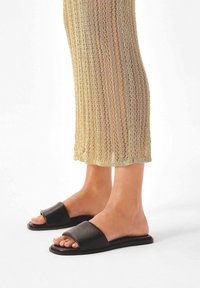 Melni ādas slidena tipa sandales ar platu siksnu, plakanām zolēm un minimālistisku dizainu. Valkātas kopā ar teksturētu zelta krāsas adītu apģērbu.