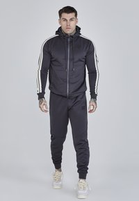 Tmavý športový teplákový súbor z mäkkého materiálu, s kapucňou na zips a bielymi bočnými pruhmi, a so zúženými nohavicami s rebrovanými manžetami.