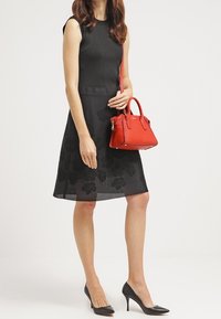 Černé bezrukávové šaty s květinovou krajkovou vsadkou, v kombinaci s malou červenou kabelkou a černými vysokými podpatky, stojící na bílém pozadí.