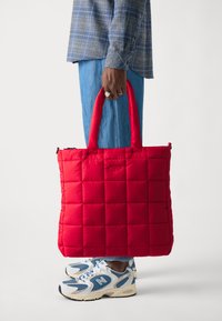 Sac fourre-tout matelassé rouge avec rembourrage, deux poignées et fermeture à glissière. Porté avec un jean en denim bleu clair et des chaussures de sport.