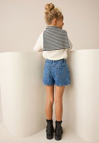 Witte blouse met lange mouwen, gestreepte zwart-witte kraagwikkel, blauwe spijkershort en zwarte enkellaarzen met grove zolen, staand tegen een muur.