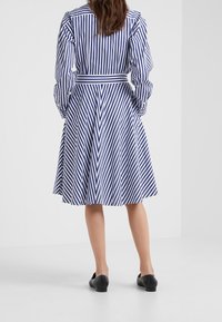 Kvinna som står vänd bort, iklädd en blå- och vitrandig skjortklänning med svarta platta skor mot en enkel vit bakgrund.