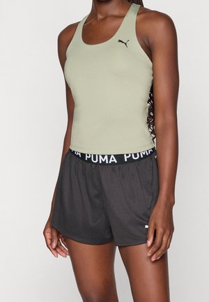 Mujer con camiseta sin mangas Puma verde claro y pantalones cortos negros Puma sobre un fondo liso.