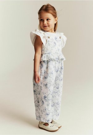 Ein junges Mädchen steht, trägt ein weißes Kleid mit Blumenmuster und Rüschenärmeln sowie cremefarbene Schuhe, schaut mit einem leichten Lächeln zur Seite.