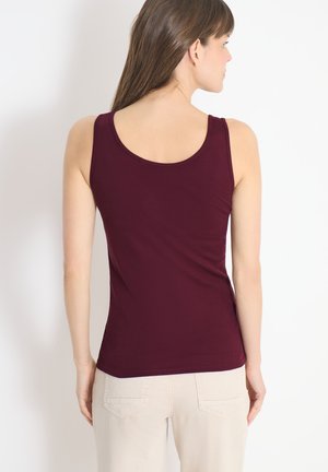 Femme aux longs cheveux bruns portant un débardeur sans manches violet foncé et un pantalon beige clair, vue de dos sur un fond blanc.