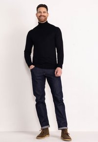 Maglione nero a collo alto, jeans blu slim-fit e stivali in suede marrone. Modello in piedi in modo informale con le mani in tasca su uno sfondo neutro.