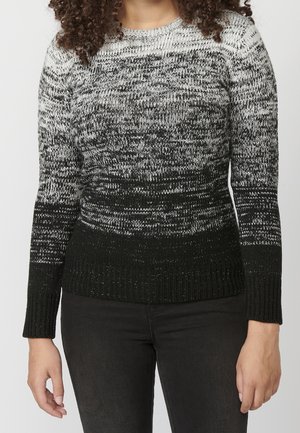 Femme portant un pull en maille dégradé noir et blanc à manches longues avec un pantalon noir, debout devant un fond clair uni.