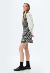 Vestido a cuadros blanco y negro con un cuerpo ajustado y falda acampanada, combinado con una blusa blanca de mangas abullonadas y mocasines negros con calcetines.