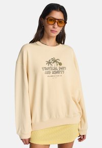 Φαρδιά φούτερ σε κρεμ απόχρωση από μαλακό μείγμα βαμβακιού, με τροπικό σχέδιο που απεικονίζει φοίνικες και κείμενο. Με ριμπ μανσέτες και τελείωμα.