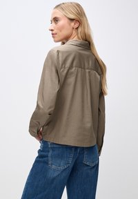 Khakifarbenes Hemd mit Knöpfen, Kragen, langen Ärmeln und einem Rückenpasse-Design. Kombiniert mit blauer Jeans, die eine lockere Passform und eine glatte Textur zeigt.