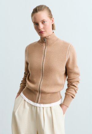 Mladá žena s blond vlasmi, oblečená v béžovom svetri na zips a krémových nohaviciach, stojaca s rukami v vreckách na svetlom pozadí.
