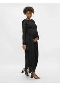 Robe de maternité avec un haut en dentelle noire à manches longues, une jupe fluide en tissu transparent et une taille froncée, accompagnée de sandales à talons noires à lanières.