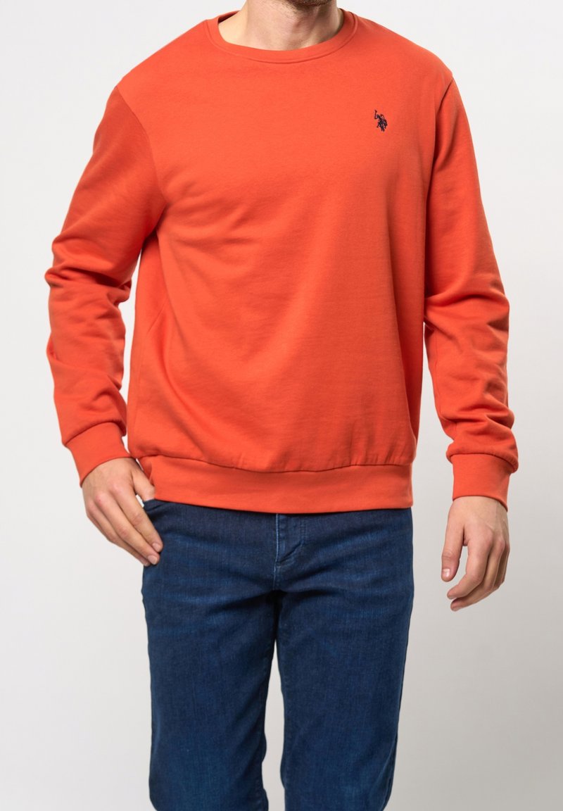 Oranger Sweatshirt aus Baumwolle mit Rundhalsausschnitt und langen Ärmeln, versehen mit einem kleinen blauen Logo auf der oberen linken Seite. Kombiniert mit blauen Jeans.
