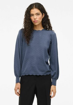Femme aux cheveux frisés et foncés, portant un haut bleu texturé à manches longues et un pantalon noir, se tenant contre un fond blanc uni.