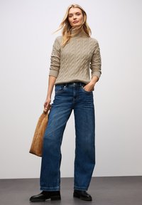 Hellbeige, kabelgestrickter Rollkragenpullover, weite, dunkle blaue Denim-Jeans, schwarze Stiefeletten und eine handtasche aus hellbraunem Wildleder. Einfaches, lässiges Outfit.
