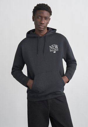 Joven con las manos en los bolsillos, vistiendo una sudadera negra de New Balance y pantalones oscuros, frente a un fondo liso y claro.