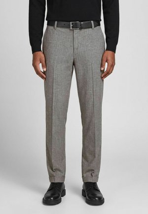Homme portant un pantalon habillé gris avec des plis à l’avant, une ceinture noire, un haut noir à manches longues et des chaussures habillées noires à lacets, debout devant un fond uni.