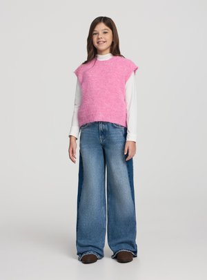 Ragazza giovane in piedi, indossa un maglione rosa senza maniche sopra una camicia bianca a maniche lunghe, jeans blu a gamba larga e scarpe marroni, sorridente.