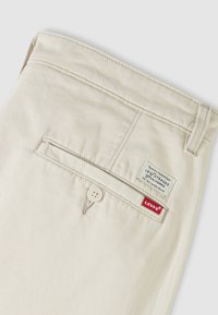 Béžové nohavice Levi's s odvetraným zadným vreckom so zapínaním na gombík a biało-červenými štítkami s logom Levi's na hornej pravej strane.