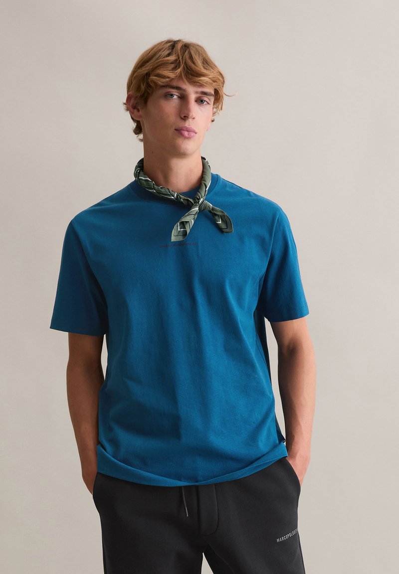 Turkusowa bawełniana koszulka z krótkimi rękawami, okrągłym dekoltem i detalem z logo. Noszona z zielonym wzorzystym szalikiem na szyję i czarnymi spodniami.