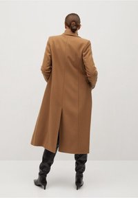 Bruine wollen lange jas met een notch kraag, zijzakken en een rugventilatie, gecombineerd met zwarte laarzen met hoge hakken tot boven de knie.