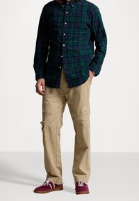 Mies, joka pitää tummanvihreää ja laivastonsinistä ruutukuviollista paitaa, beigejä kuluneita housuja ja bordoynhuoneisia tennareita, joissa on valkoiset nauhat ja raidat.