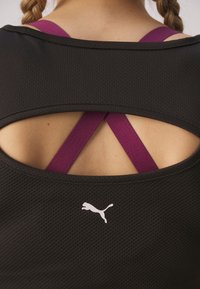 Črna športna majica s teksturiranim zaključkom, z globokim izrezom na hrbtni strani in križanimi vijoličastimi naramnicami. Bela logo na spodnjem srednjem delu.